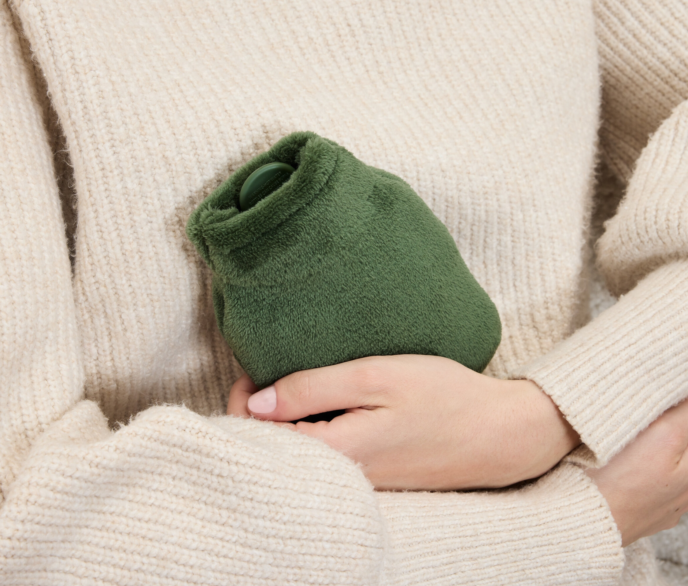 Žena v svetru drží v rukou zelenou plyšovou termoforovou láhev.