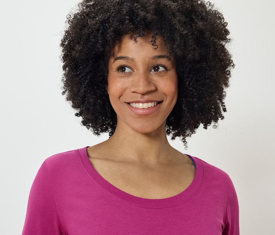 Portrét usmívající se ženy s afro účesem v růžovém tričku, dívající se doleva.