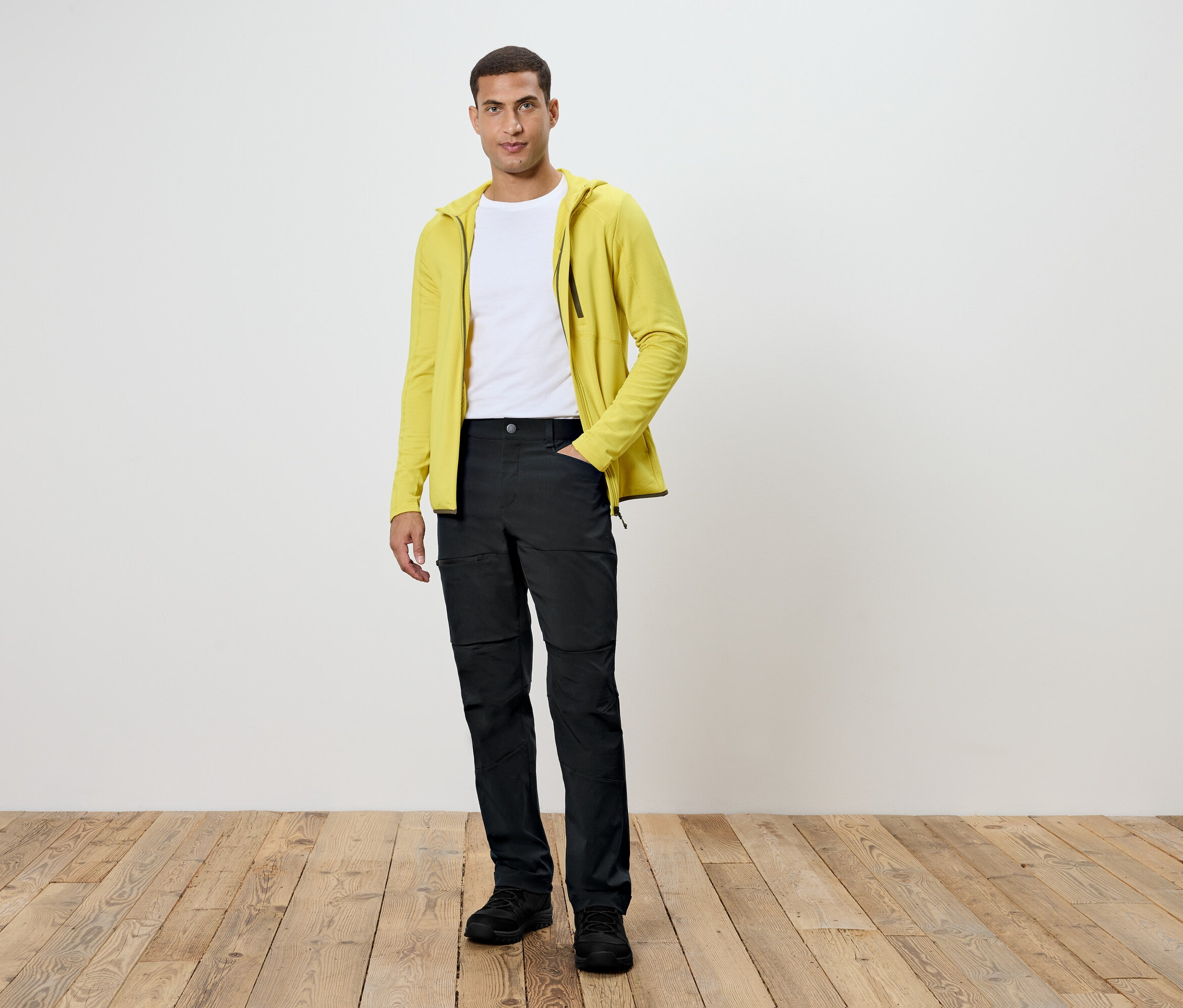 Muž stojí na dřevěné podlaze. Má na sobě černé kalhoty, bílé tričko a žlutou mikinu.