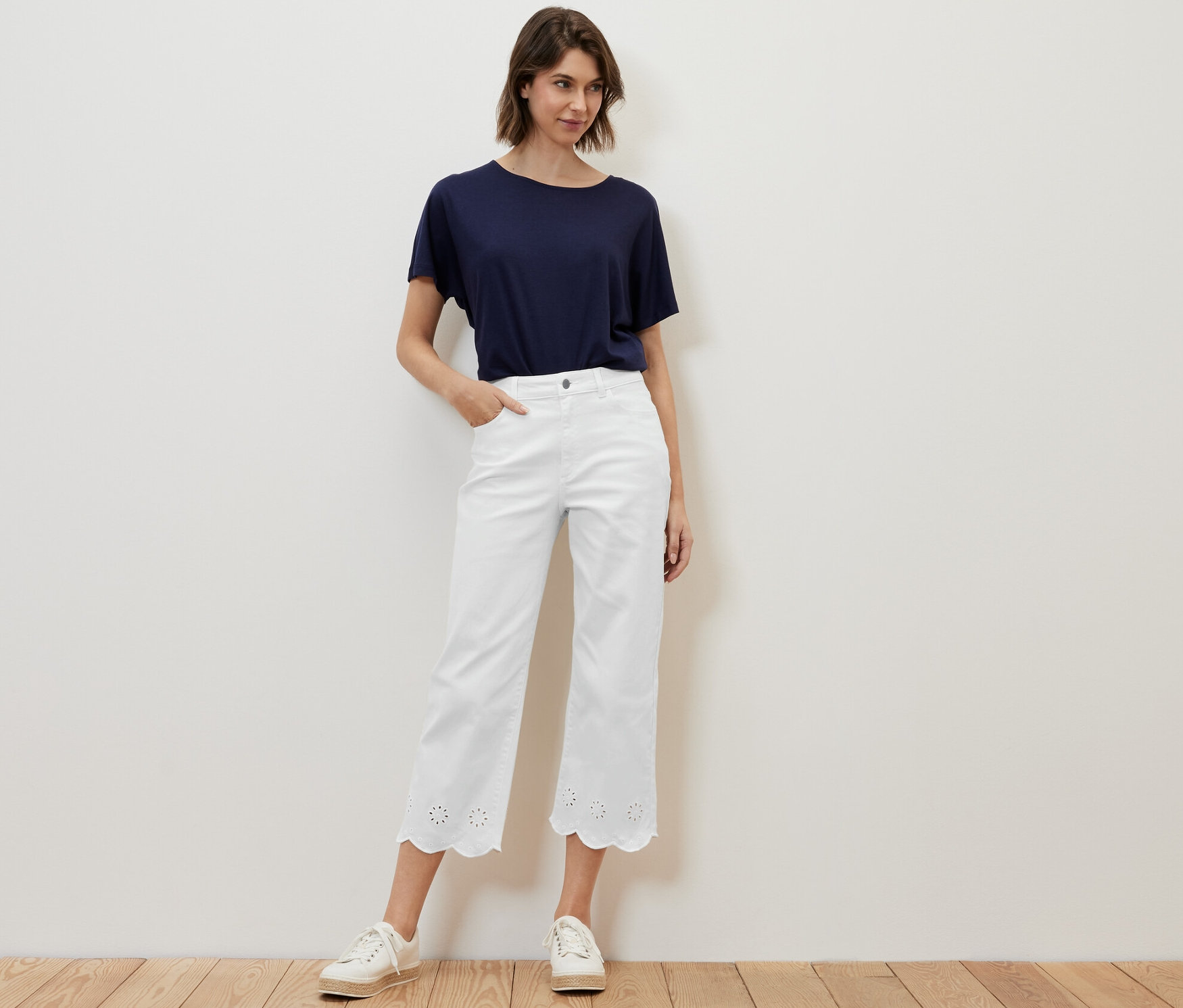 Žena pózuje v bílých džínách s ažurovou výšivkou a modrém tričku.