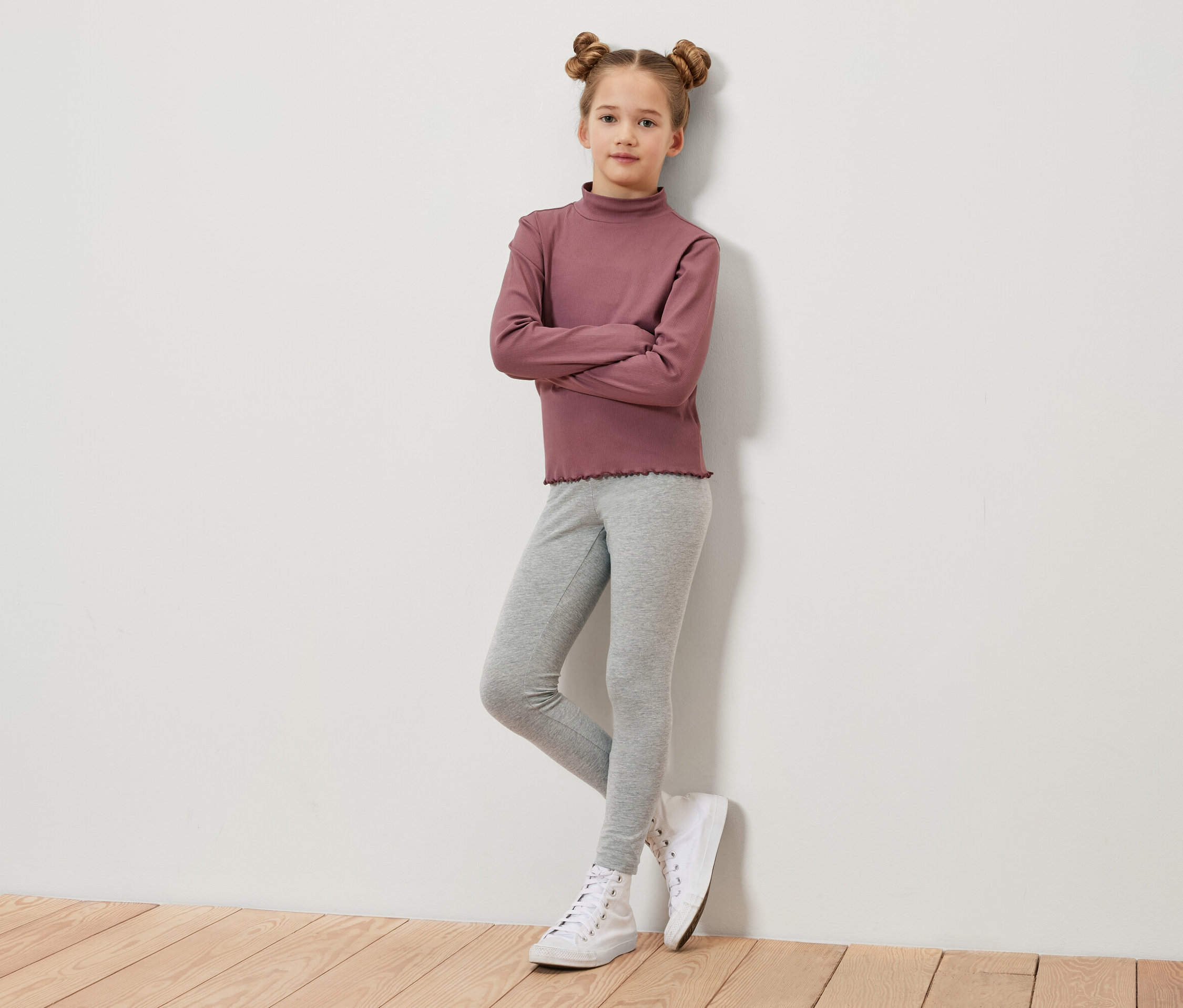 Dívka s hnědými vlasy sepnutými do drdolů stojí s rukama zkříženýma na hrudi. Má na sobě vínové tričko s dlouhým rukávem, šedé legíny a bílé tenisky.