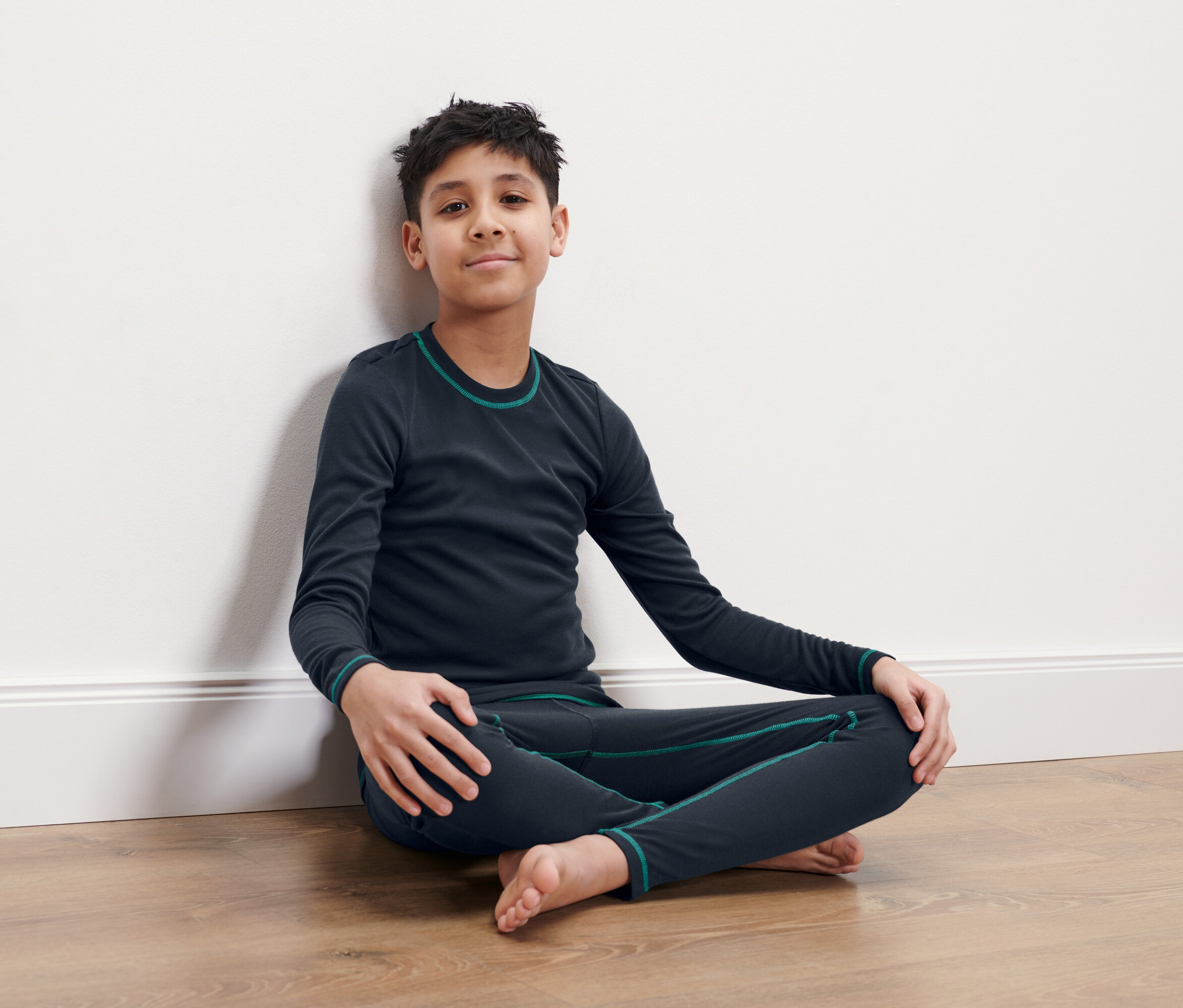 Chlapec sedí se zkříženýma nohama na podlaze a opírá se o bílou stěnu. Má na sobě tmavě modré termoprádlo.