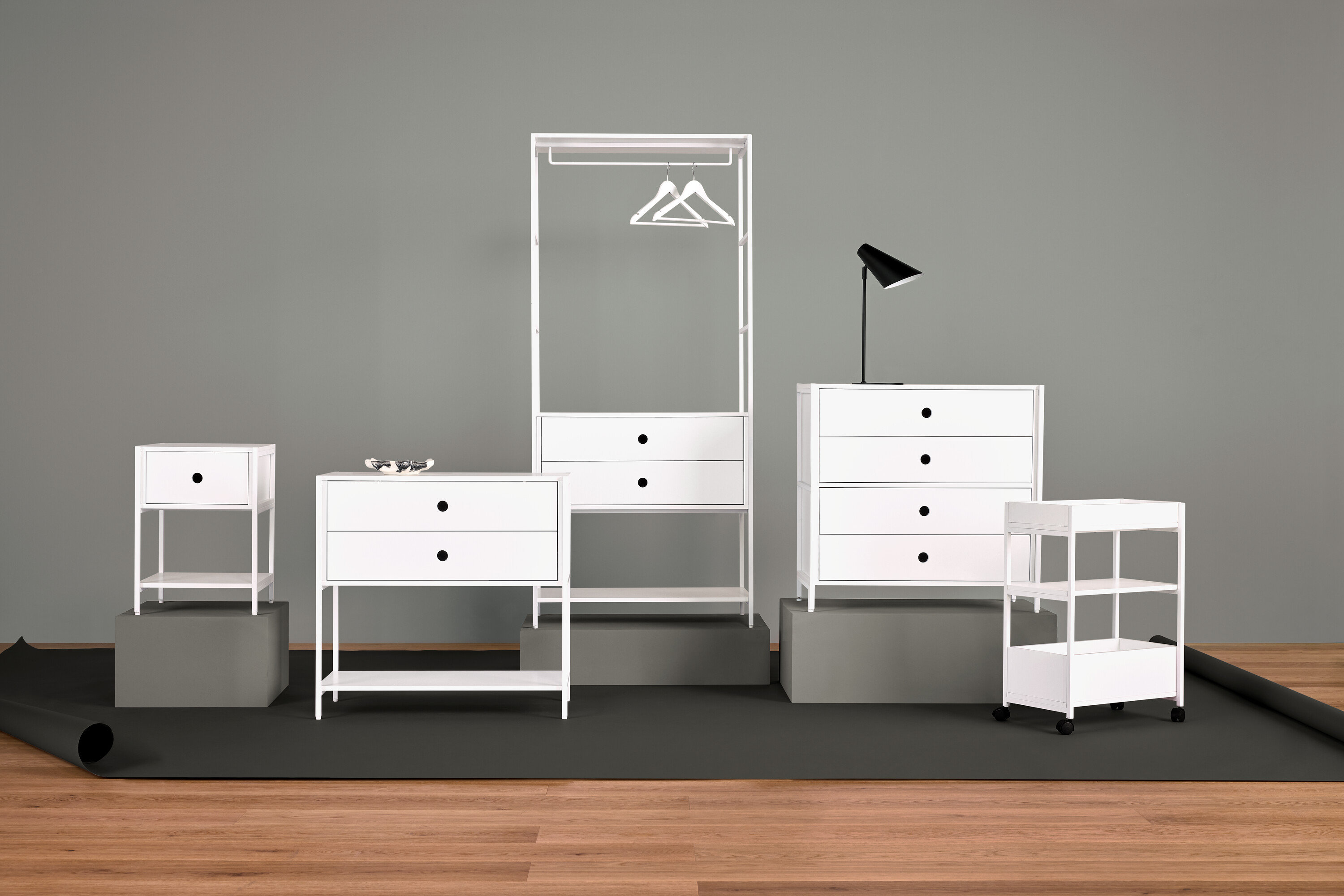 Sestava bílého nábytku s nočním stolkem, komodou se zásuvkami, šatním věšákem, lampou a policemi.