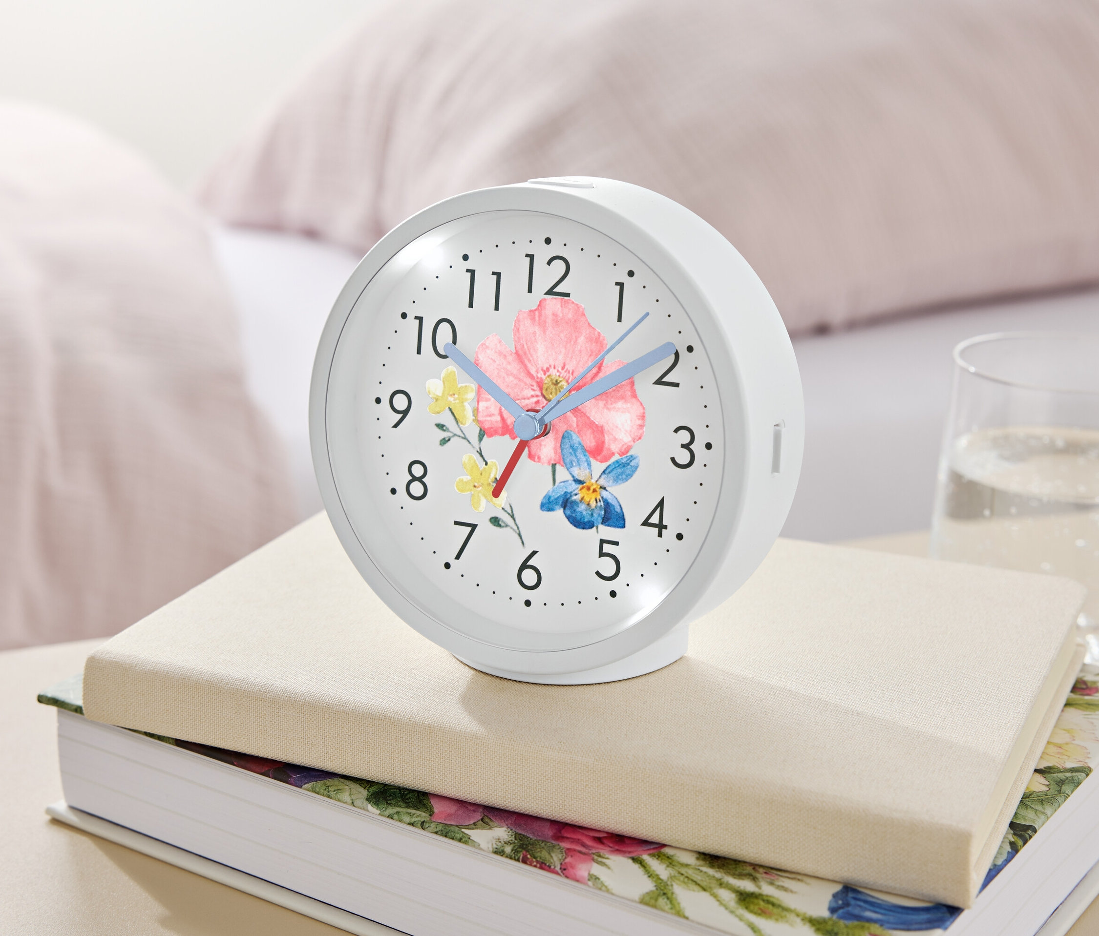 Bílý budík s květinovým motivem leží na knihách vedle sklenice vody.