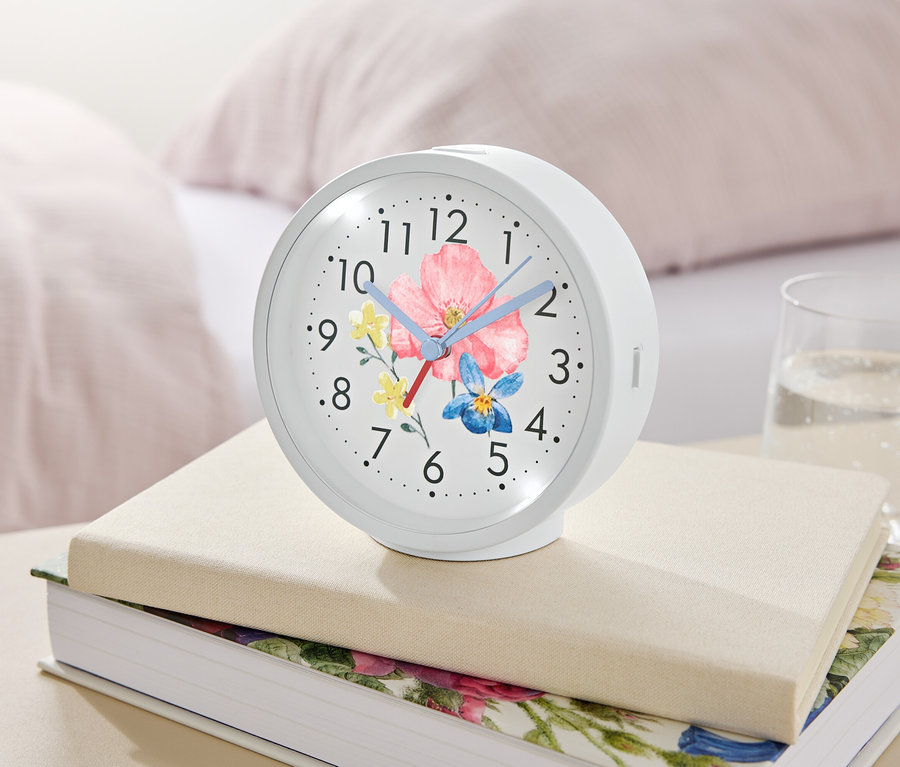 Bílý budík s květinovým motivem leží na knihách vedle sklenice vody.