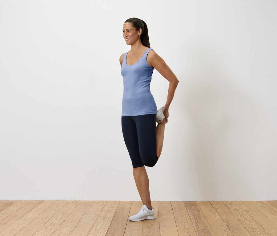 Žena v modrém tílku a tmavých legínách si protahuje kvadriceps držením nohy za zády.