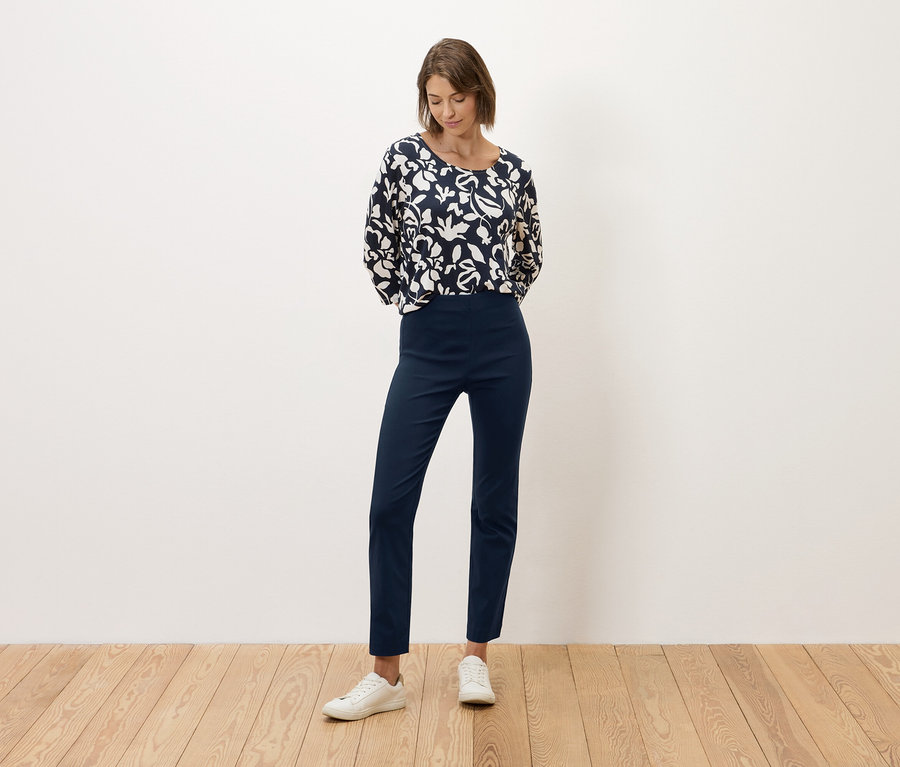 Žena stojí před bílou zdí, oblečená do tmavě modrých kalhot a halenky s černobílým vzorem.