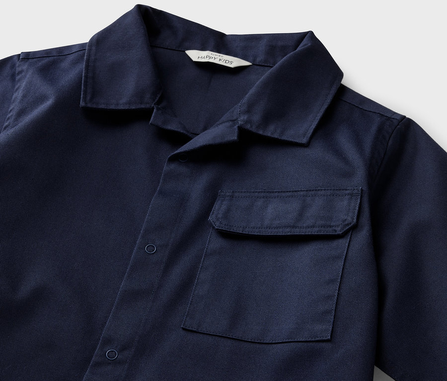 Detail modré dětské svrchní košile s krátkým rukávem a kapsou.