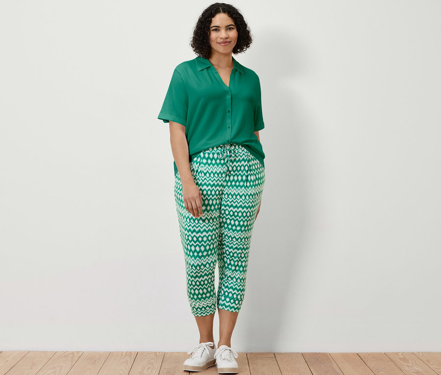 Žena stojí a má na sobě smaragdově zelené halenkové triko s knoflíkovou légou a 3/4 žerzejové kalhoty s potiskem.