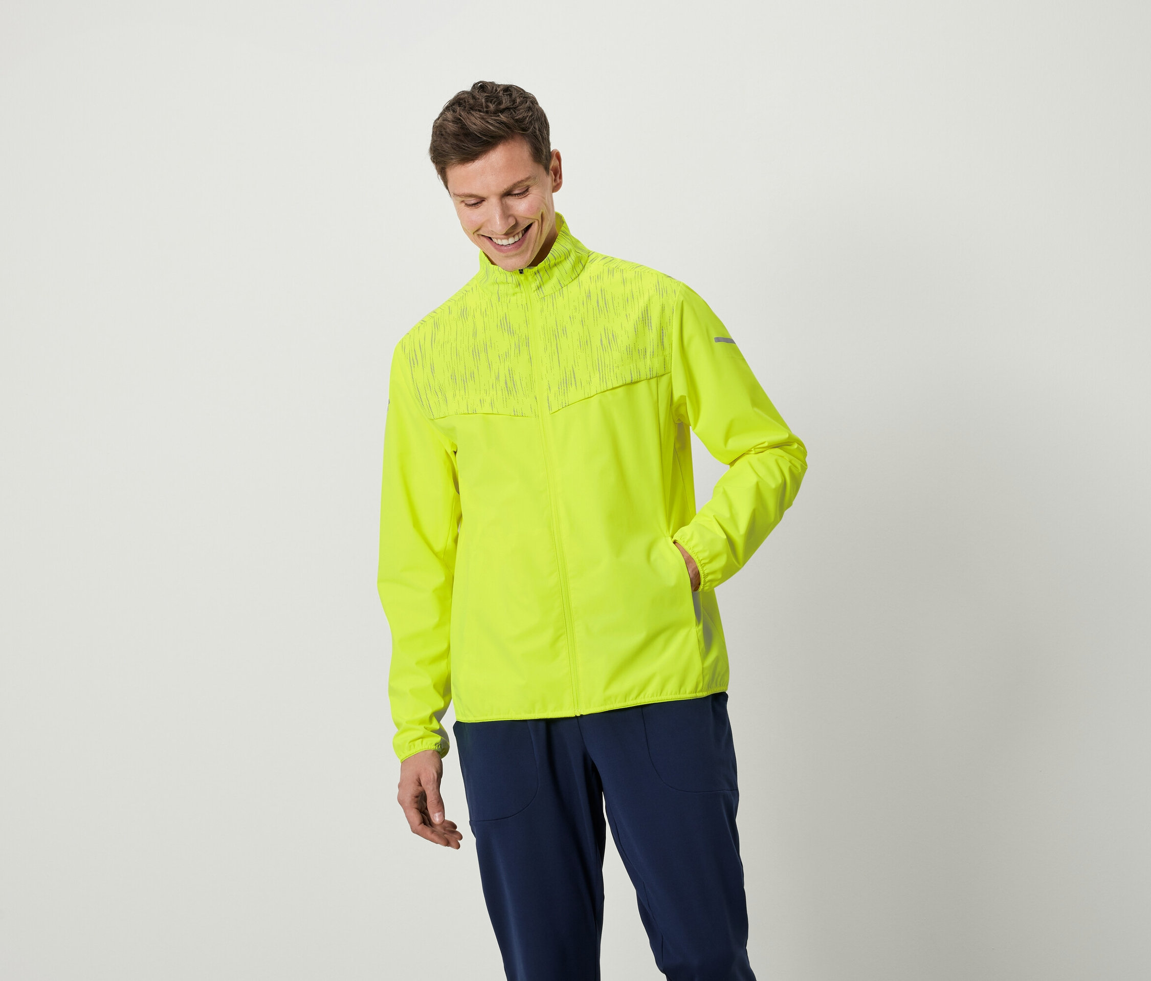 Muž se usmívá a pózuje v neonově žluté bundě a tmavě modrých kalhotách.
