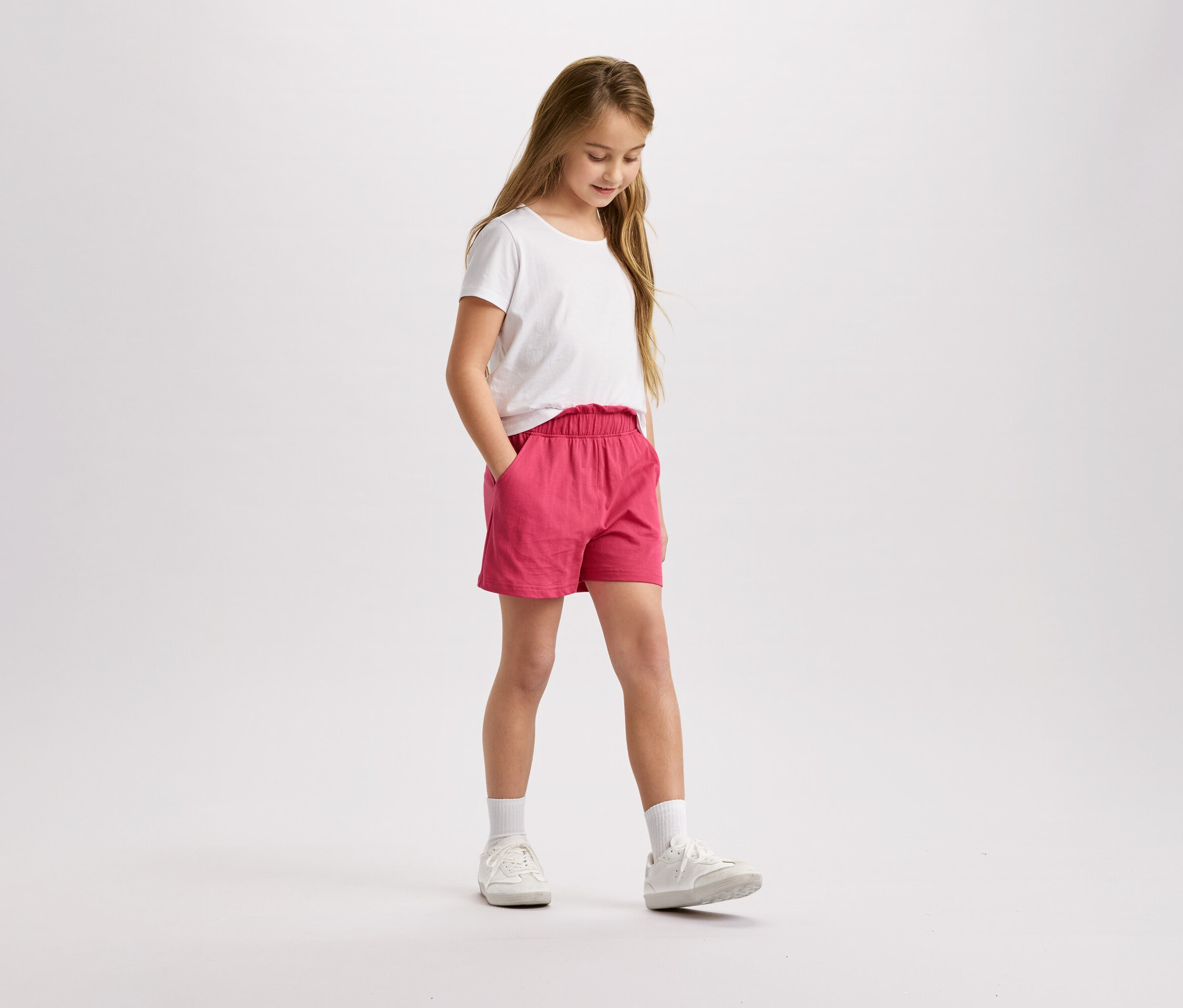 Dívka s blond vlasy v bílém tričku a růžových šortkách stojí a dívá se dolů.
