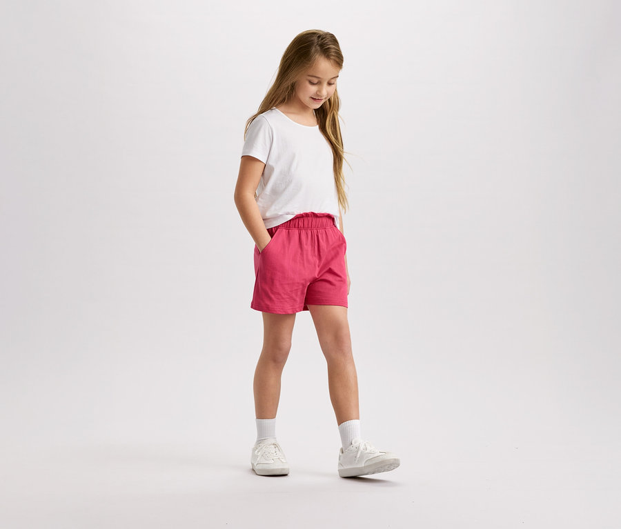 Dívka s blond vlasy v bílém tričku a růžových šortkách stojí a dívá se dolů.