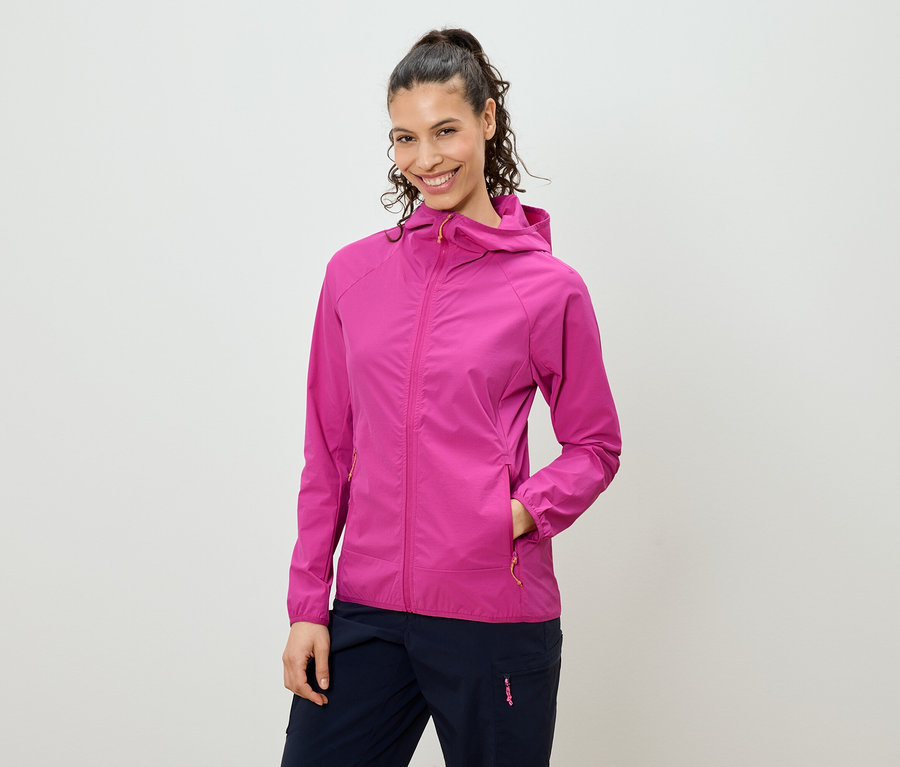 Usmívající se žena s vlasy staženými do culíku má na sobě růžovou bundu s kapucí a tmavě modré kalhoty.