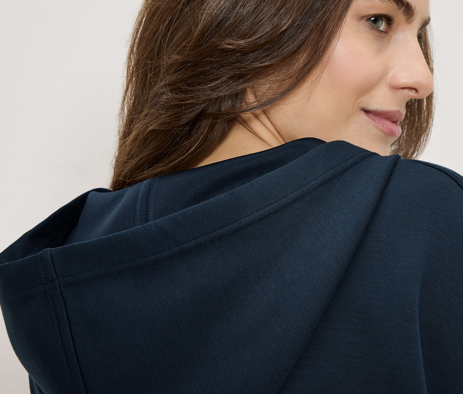 Žena má na sobě tmavě modrou mikinu s kapucí, dívá se přes rameno.