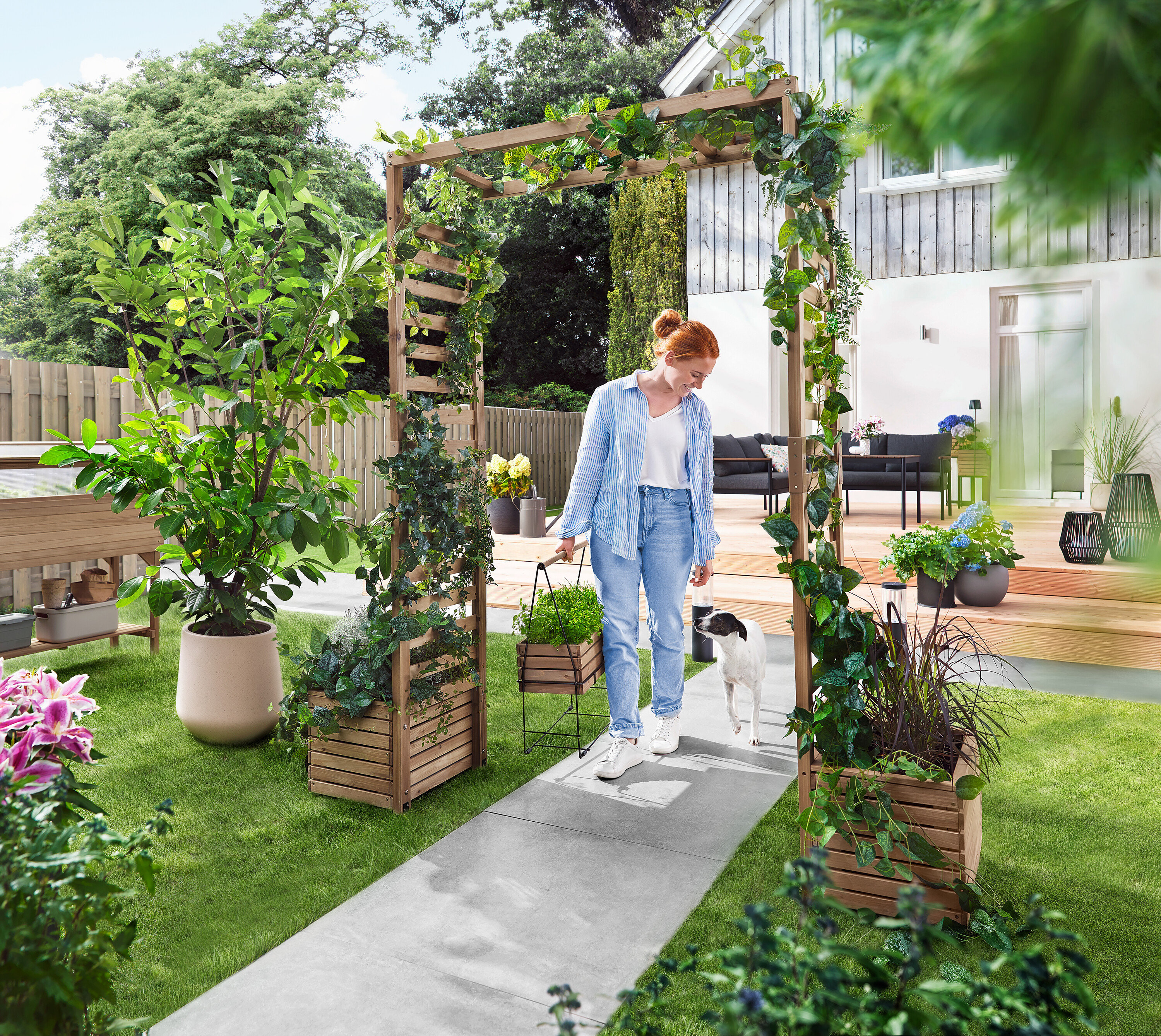 Žena prochází se psem zahradním obloukem porostlým rostlinami. Nese květináč.