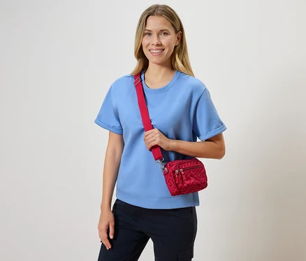 Usmívající se blondýna v světle modrém tričku a červené crossbody kabelce.