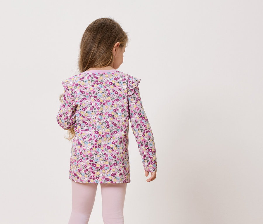 Pohled zezadu na malou holčičku s dlouhými vlasy v halence s dlouhým rukávem s květinovým potiskem a růžových legínách.