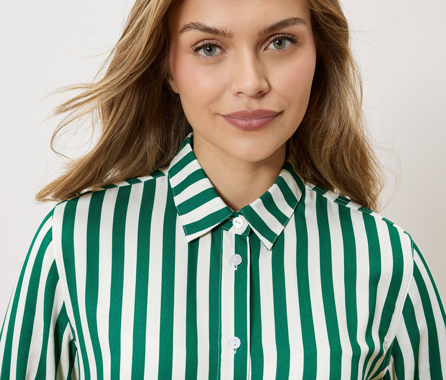 Portrét ženy s blond, vlnitými vlasy, oblečené do pruhované košile smaragdově zelené a bílé barvy.