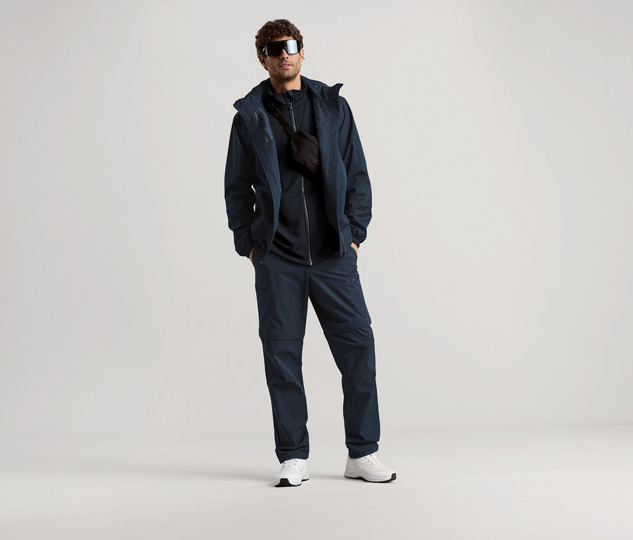 Muž stojí se slunečními brýlemi, tmavě modrou bundou, kalhotami a ledvinkou na světlém pozadí.