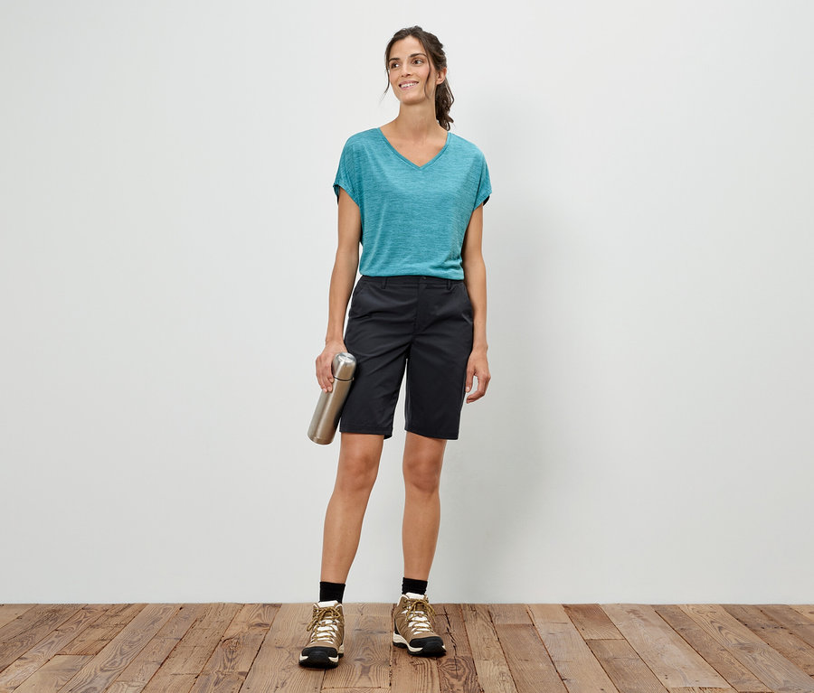 Žena stojí před bílou stěnou, drží stříbrnou láhev, má na sobě modré tričko, šortky a trekové boty.