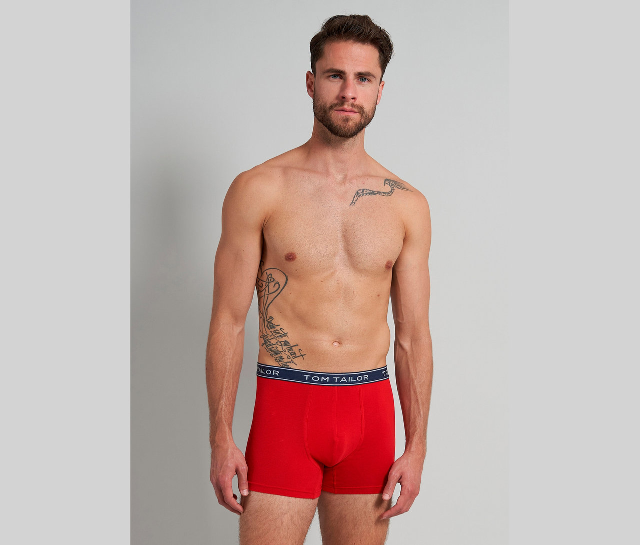 Muž s tetováním má na sobě červené boxerky Tom Tailor na šedém pozadí.