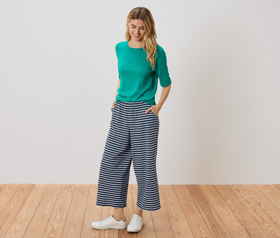 Žena stojí s rukama v kapsách v modro-bílých pruhovaných culottes, má na sobě zelený top a bílé tenisky.