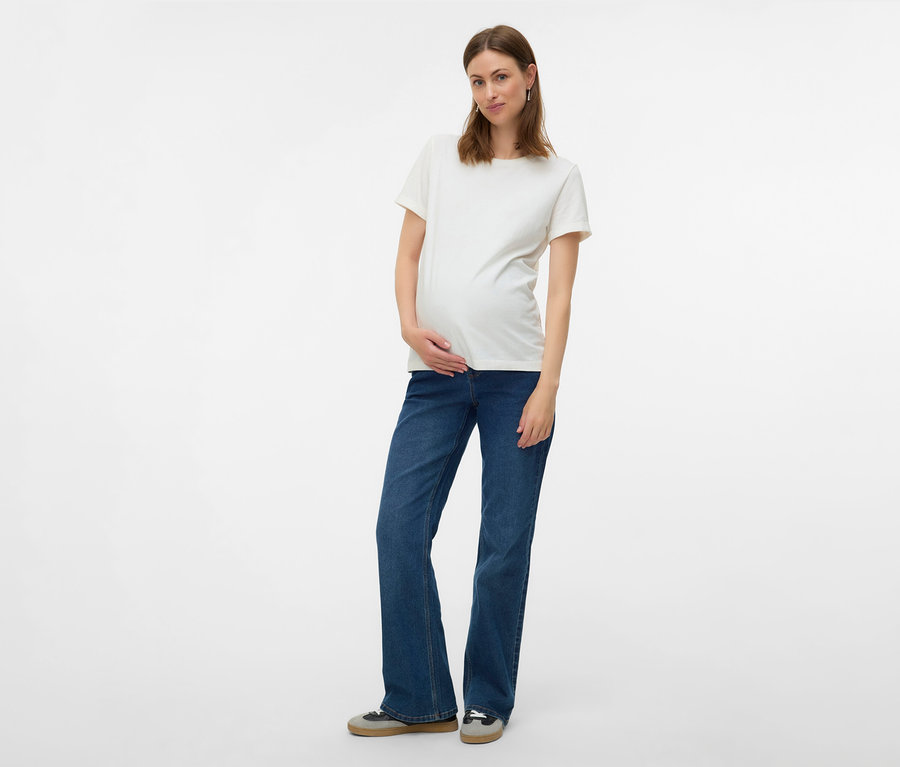 Těhotná žena v bílém tričku a džíny flared MAMALICIOUS »MLEDINA« pózuje na bílém pozadí.
