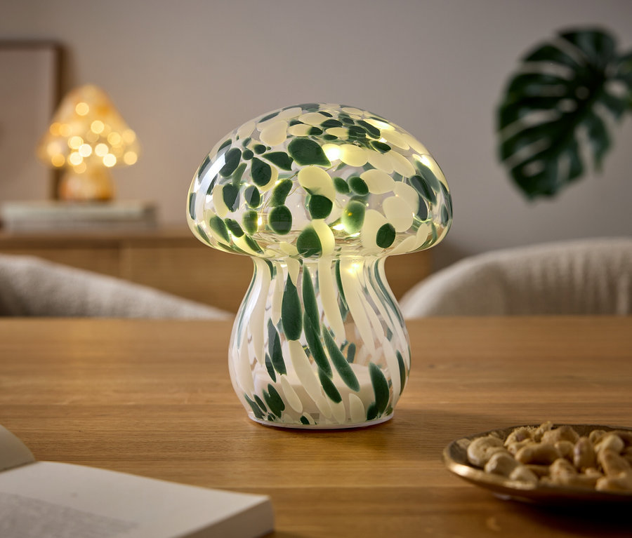 Lampa ve tvaru houby se zeleno-bílým vzorem a světly stojí na dřevěném stole. Vedle ní je otevřená kniha a malá miska s kešu oříšky. V pozadí je vidět další houbová lampa a rostlina.