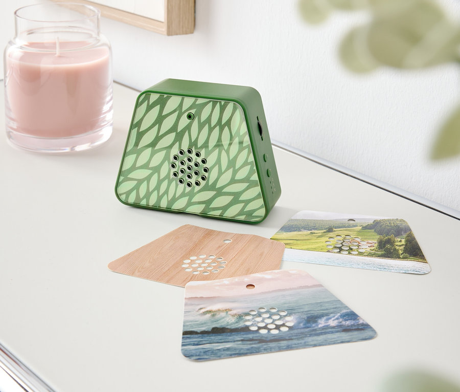 Na stole stojí růžová svíčka, zelený vonný difuzér se vzorem listů a tři vonné karty.