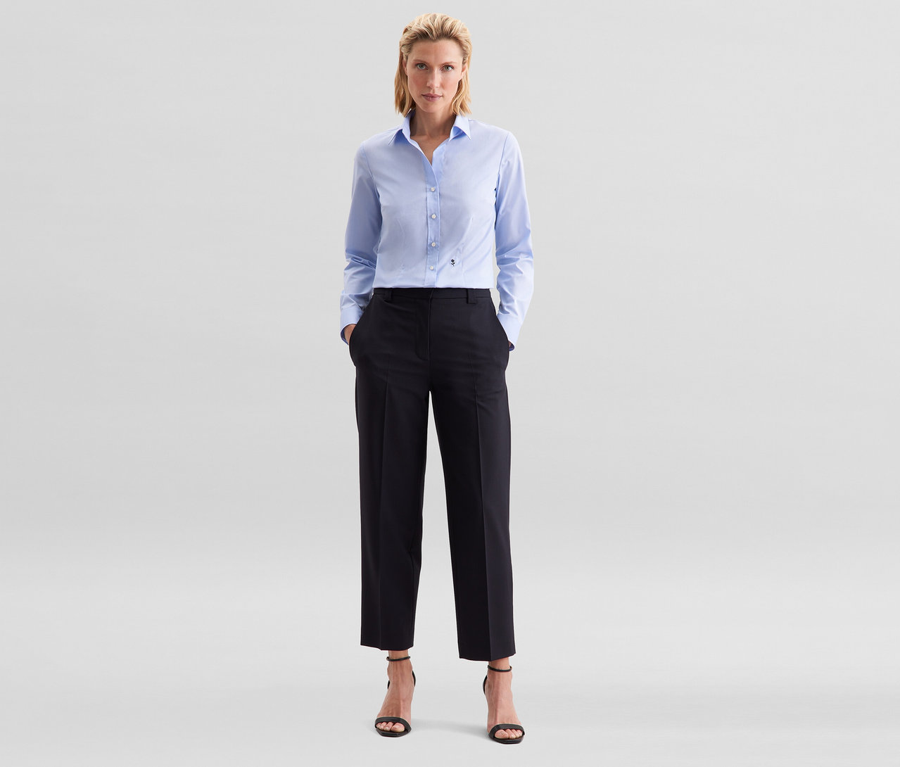 Žena stojí s rukama v kapsách, má na sobě světle modrou halenku slim fit non iron Seidensticker, černé kalhoty a černé sandály na podpatku.