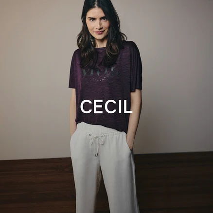 Žena s tmavými vlasy má na sobě fialové tričko s nápisem a světlé kalhoty se stahovací šňůrkou. Stojí s rukama v kapsách a dívá se do kamery.