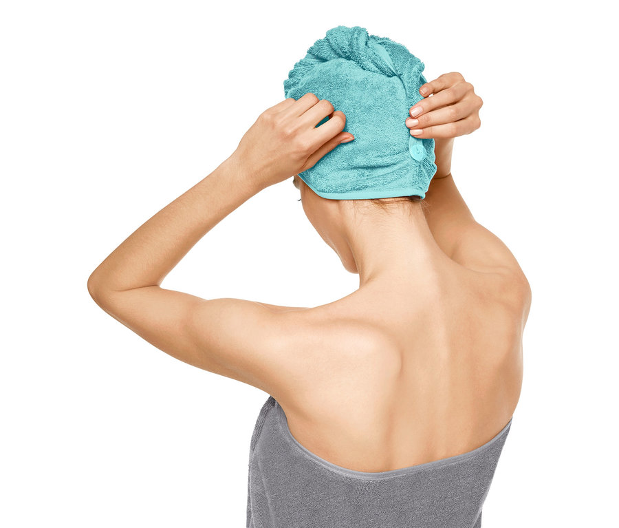 Žena si upravuje ručníkový turban na hlavě, zabalená do šedé osušky.