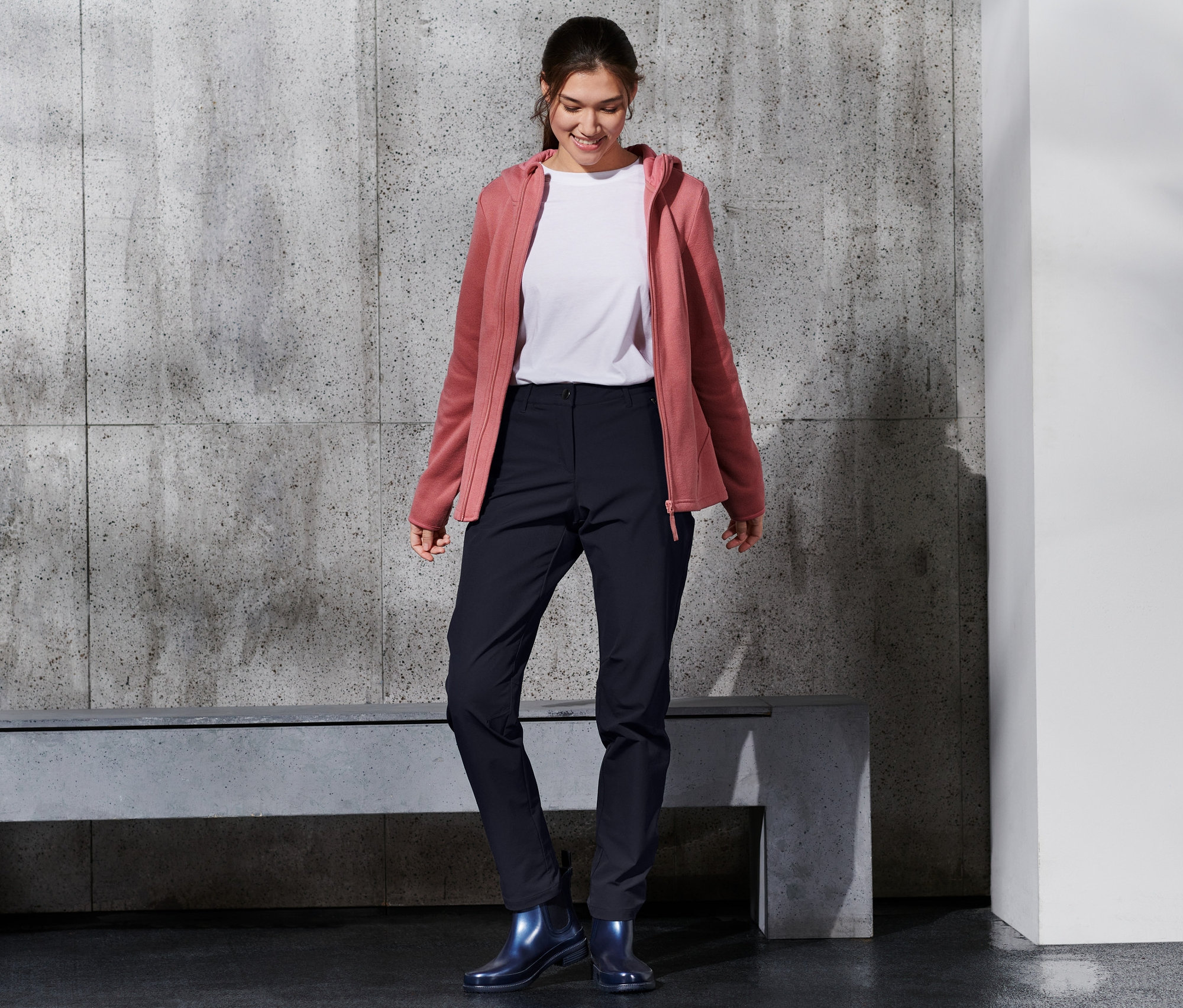 Usmívající se žena má na sobě softshellové kalhoty, bílé tričko a růžovou mikinu s kapucí.