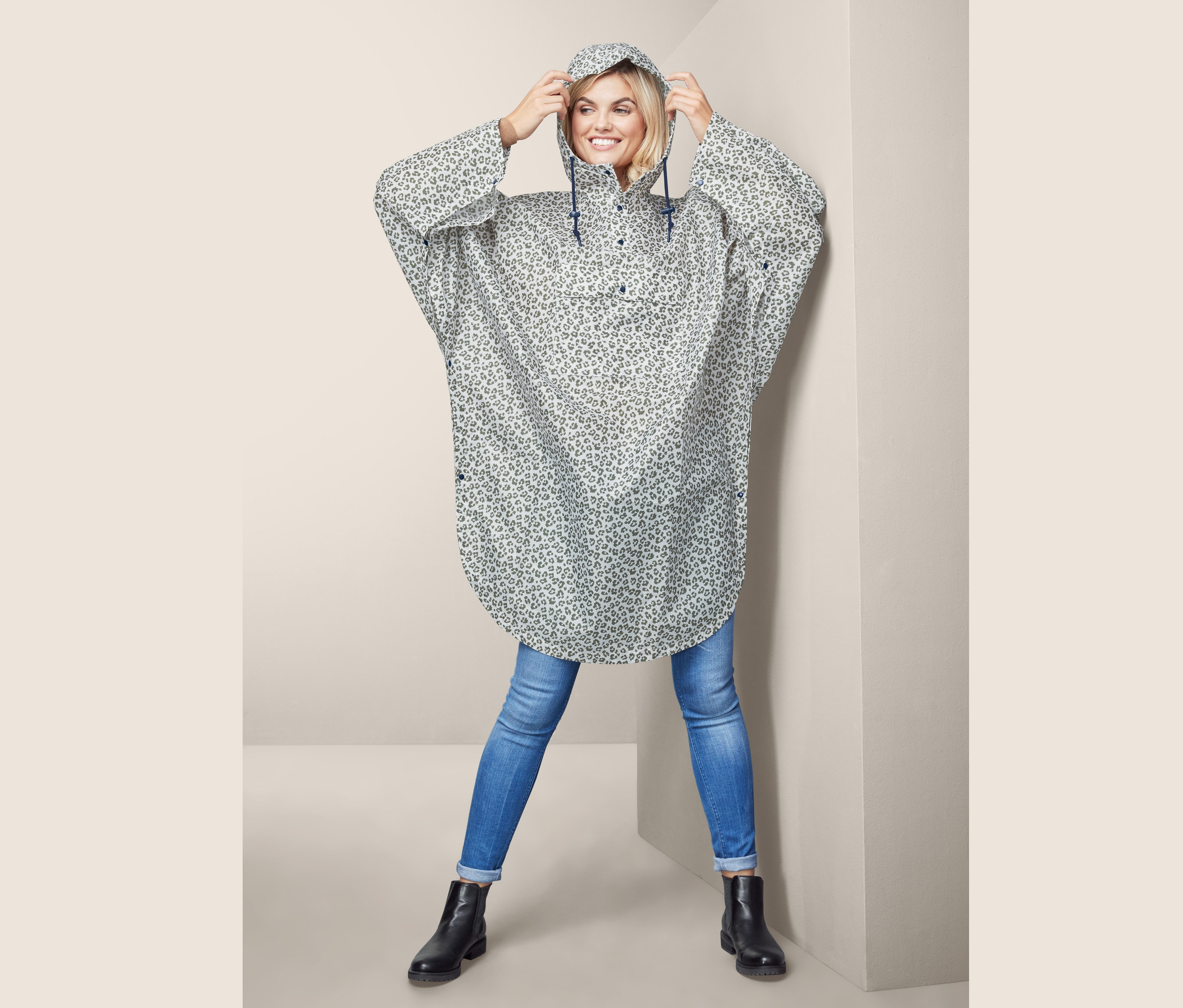 Žena si upravuje kapuci ponča do deště s leopardím vzorem, které má na sobě. Má na sobě džíny a černé boty.