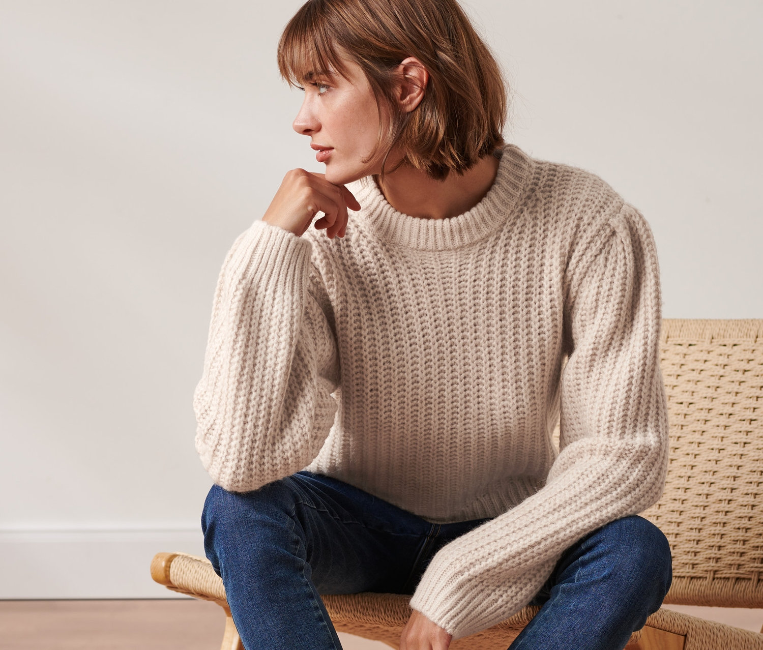 Žena sedí na židli a má na sobě krémový svetr z hrubé pleteniny s vlnou, modré džíny a černé boty.