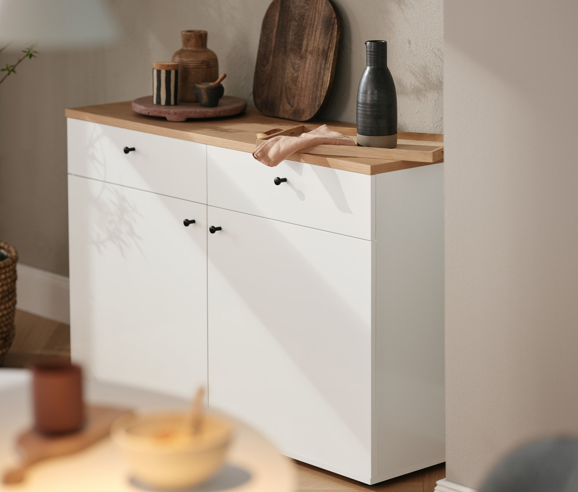 Na horní části bílé komody stojí hnědé dřevěné prkénko a různé dekorativní předměty. Nad komodou visí závěsné svítidlo Strap 16.