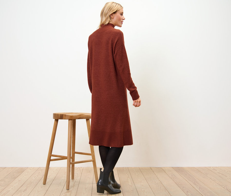 Žena stojí bokem a má na sobě pletené šaty s vlnou, černé punčochy a boty. Vedle ní stojí dřevěná stolička.