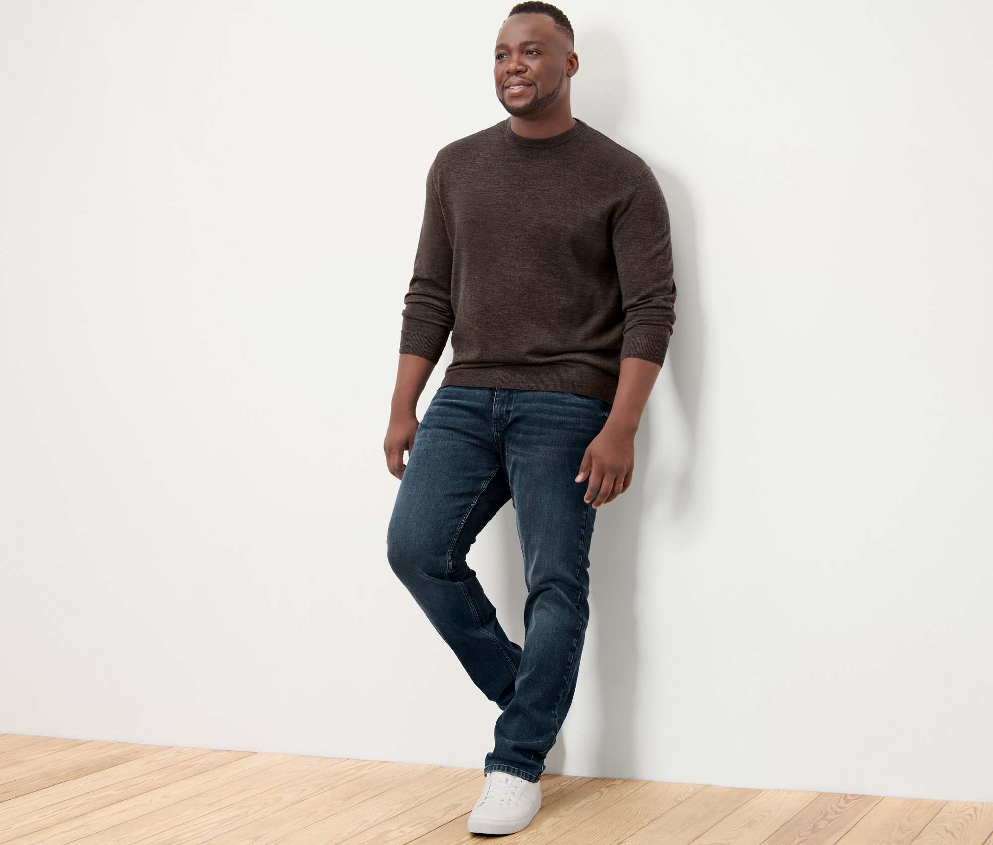 Muž opřený o zeď má na sobě hnědý svetr z merino vlny, džíny a tenisky.