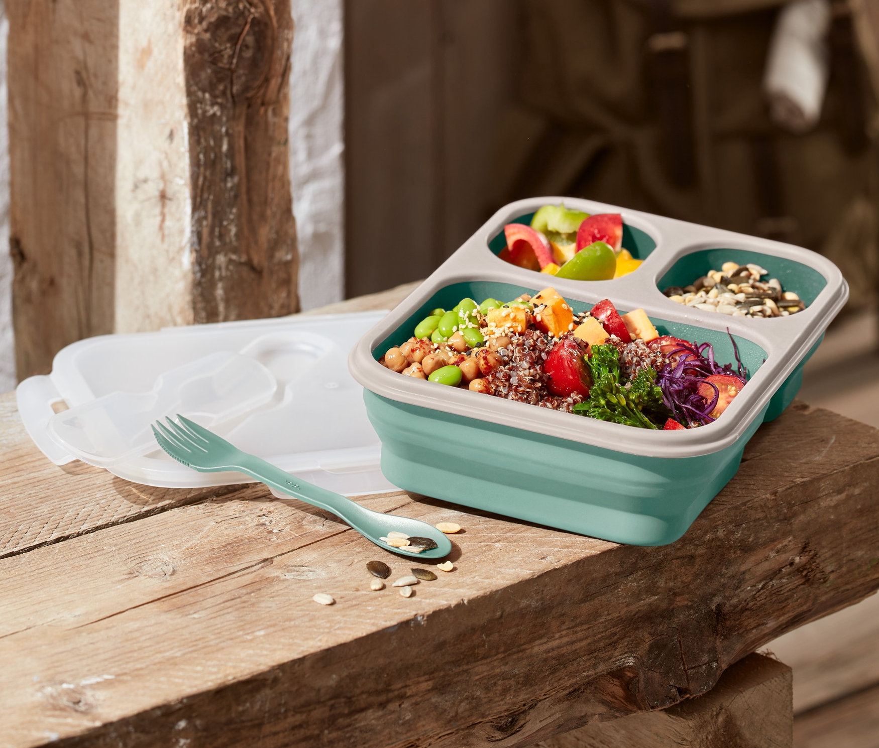 Skládací dóza na svačinu položená na dřevěné lavici, vedle je vidlička a víko.