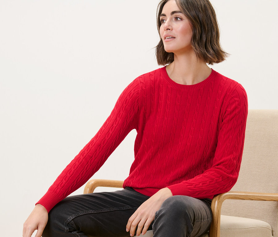 Žena sedí v křesle a má na sobě červený svetr s copánkovým vzorem.