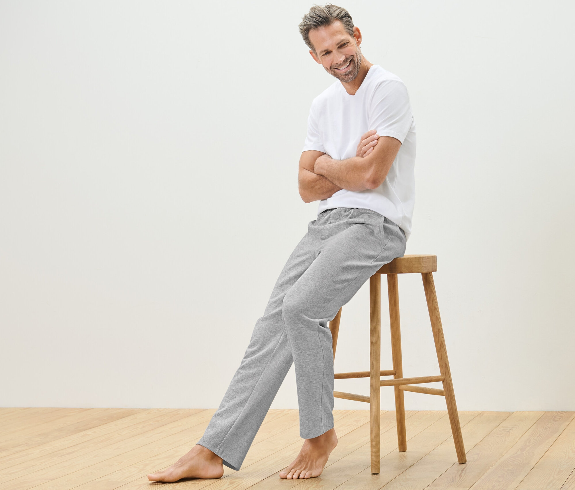 Muž sedí na dřevěné stoličce a má na sobě domácí kalhoty a tričko.