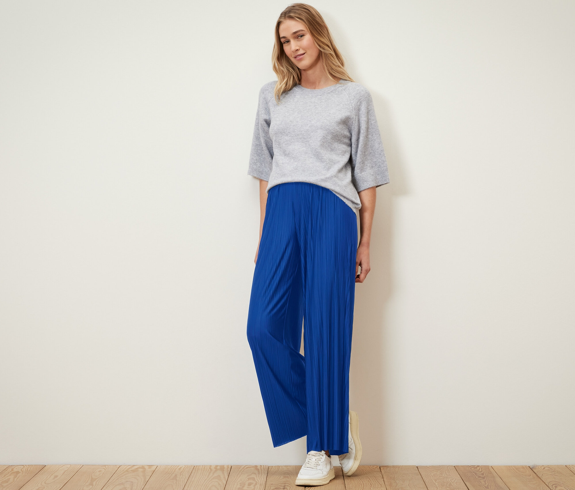 Žena stojí a má na sobě plisované kalhoty kobaltově modré barvy a šedý top.