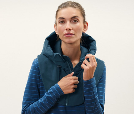 Žena si upravuje vatovanou kapuci se stojáčkem, má na sobě modré pruhované tričko a černé kalhoty.