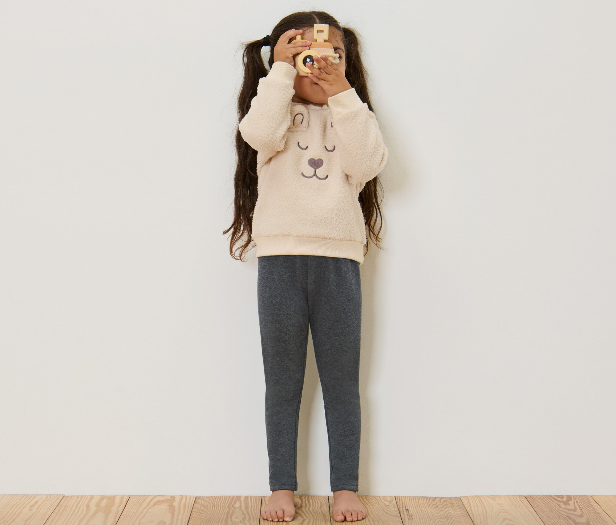 Dívka se dívá přes dřevěný fotoaparát, má na sobě šedé dětské termo legíny.