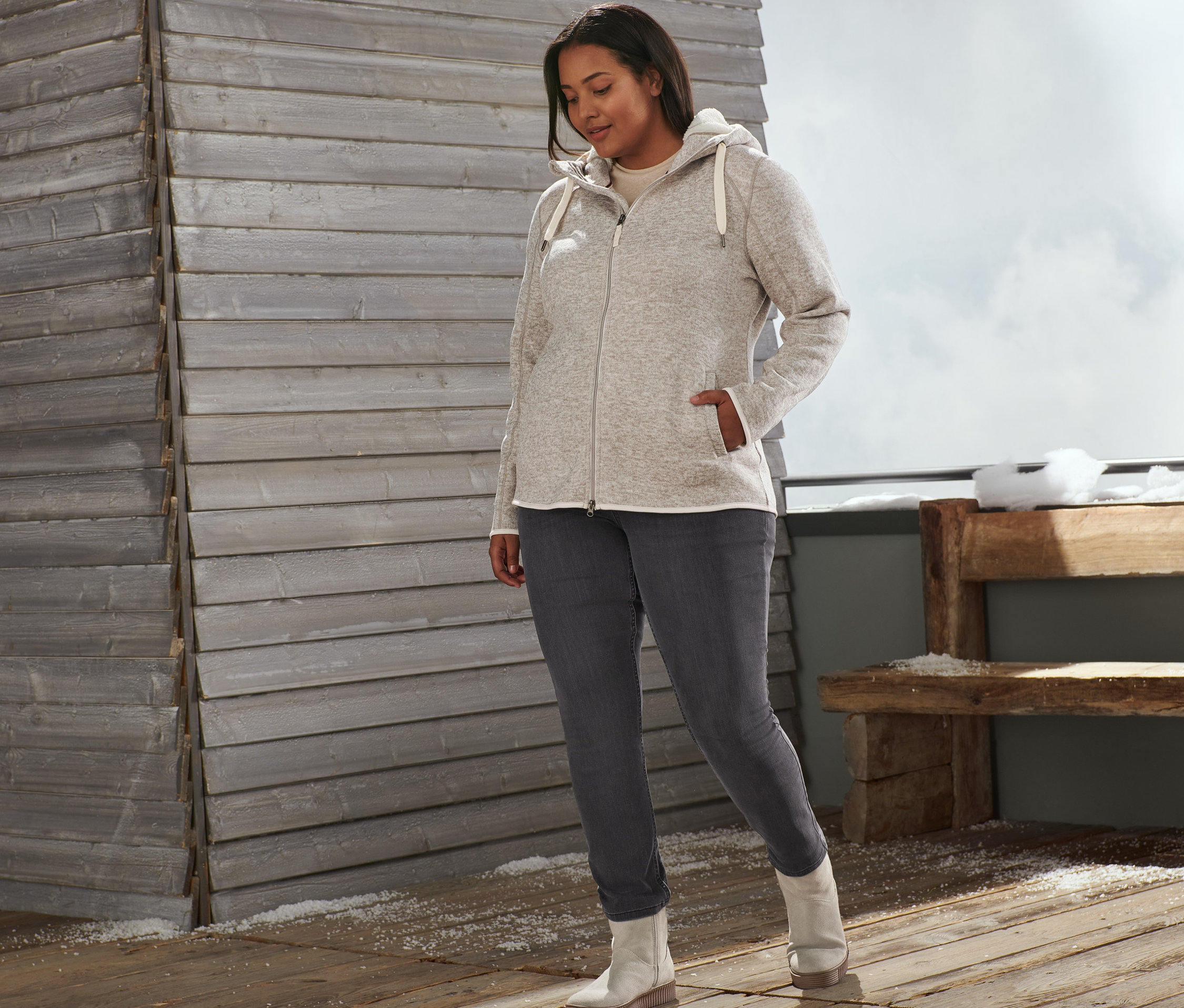 Žena stojí venku a má na sobě béžovou bundu z pleteného fleecu, džíny slim fit a kotníkové boty z velurové kůže.