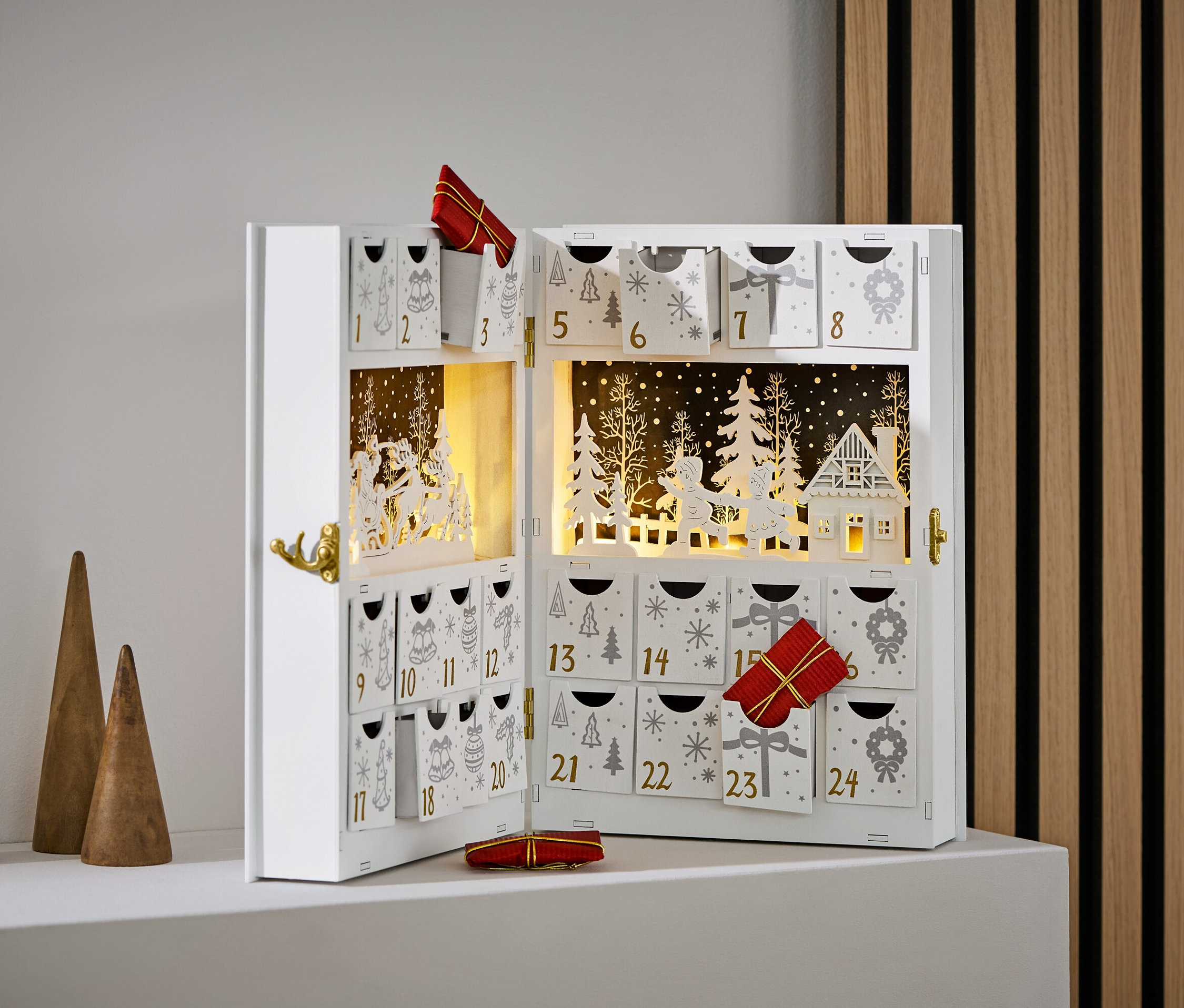 Otevřený bílý adventní kalendář s ozdobami a dárky. Stojí vedle tří dřevěných kuželů.