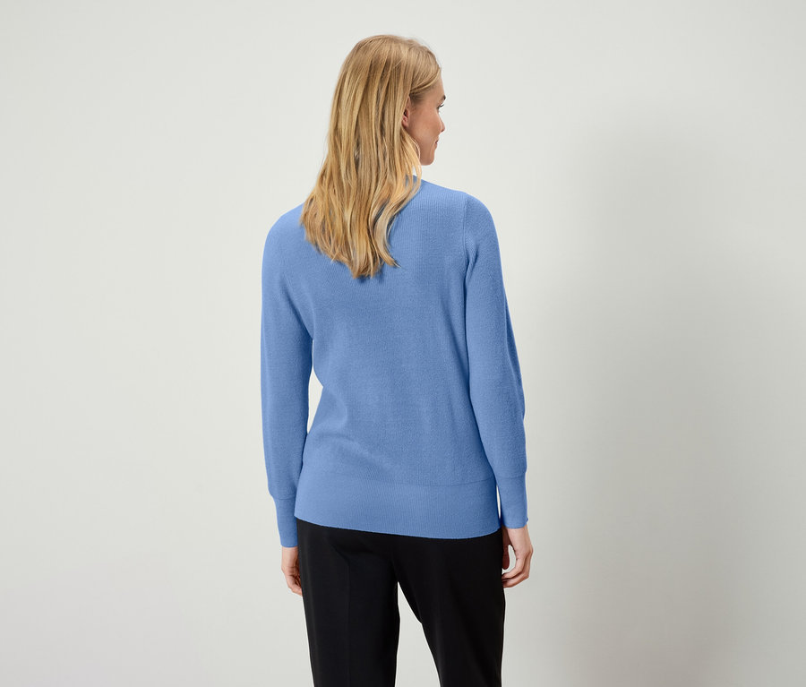 Žena v modrém svetru a černých kalhotách stojí zády k fotoaparátu.