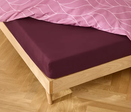 Čelo postele s bordó prostěradlem a růžovým vzorovaným povlečením.