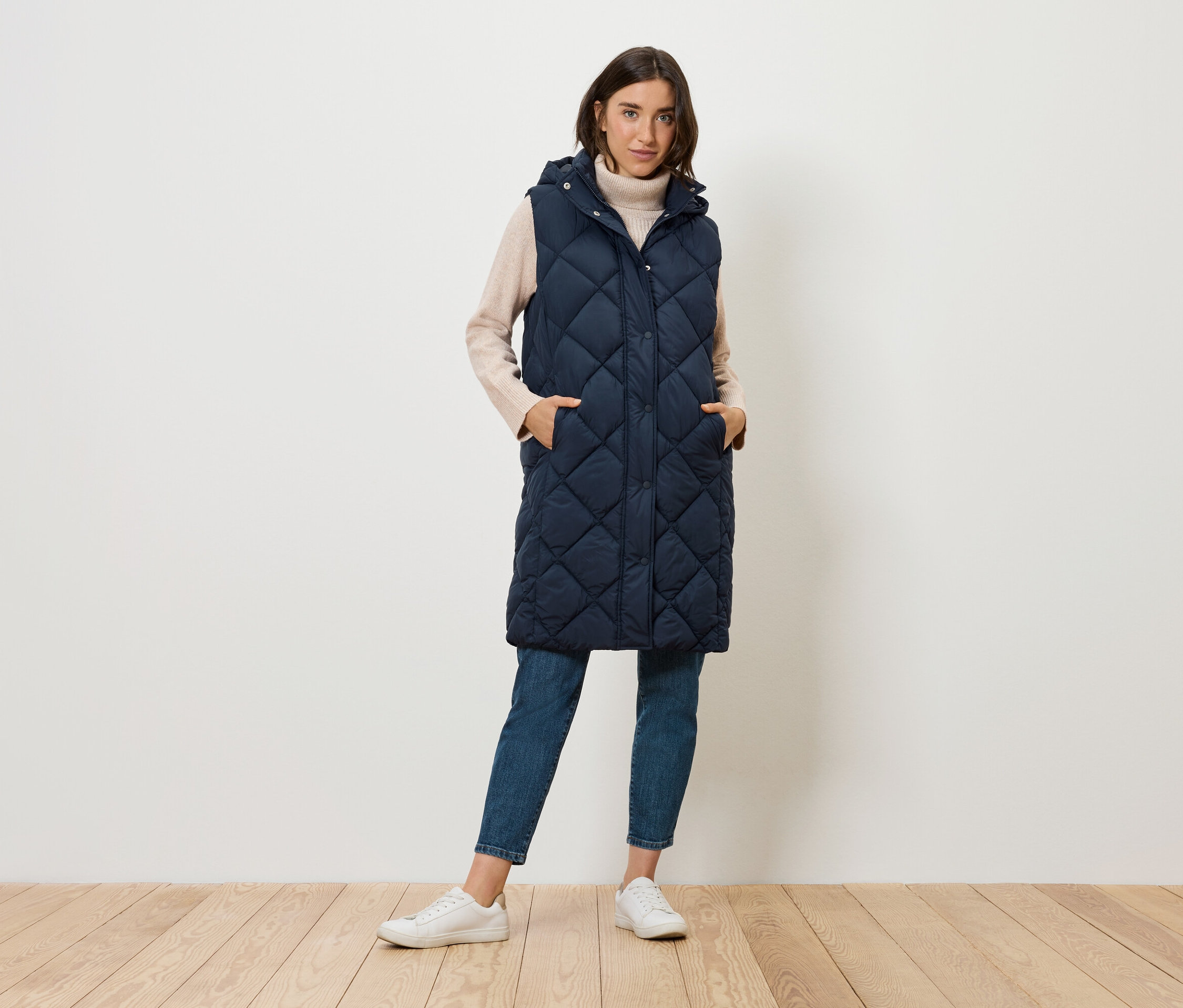 Žena stojí a má na sobě dlouhou prošívanou tmavě modrou vestu s kapucí, džíny a tenisky.