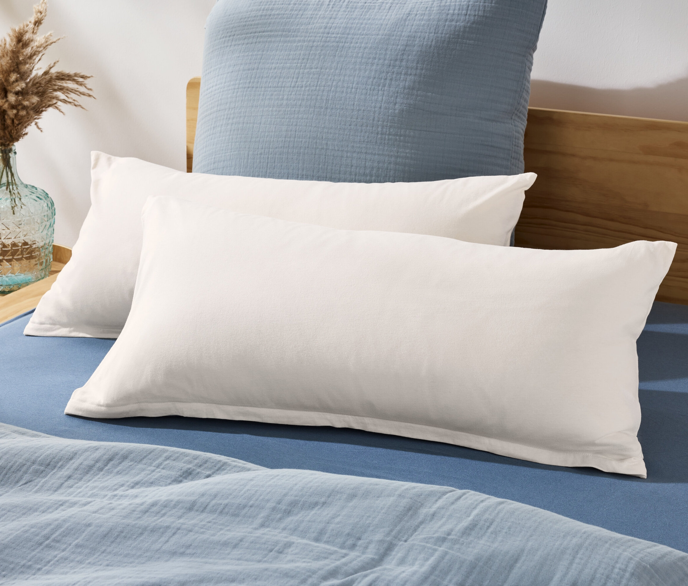 Dva žerzejové povlaky na polštáře leží na posteli s modrým prostěradlem a přikrývkou.